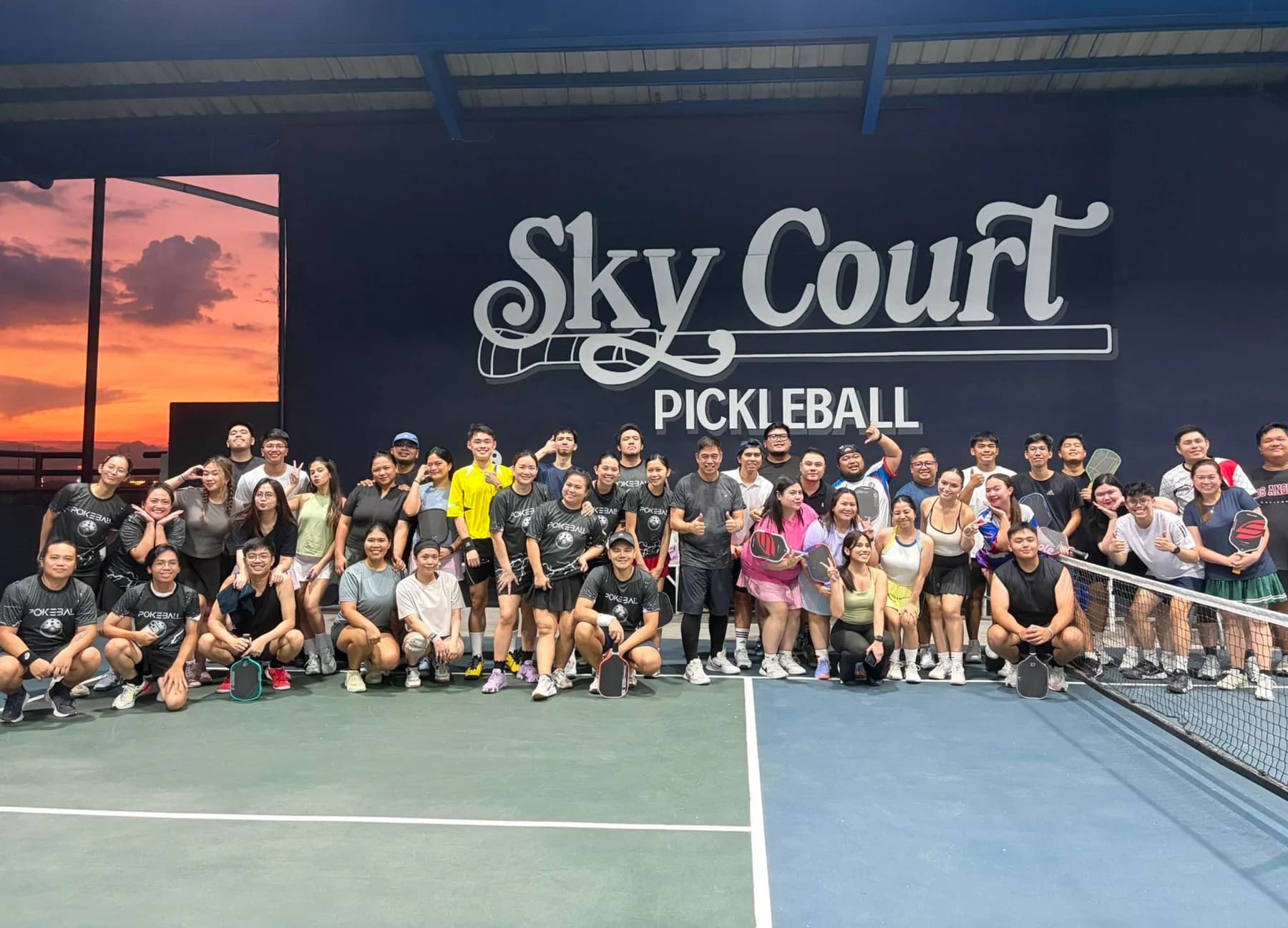 A large group of SkyCourt players pose together on court beneath the venue logo at sunset.