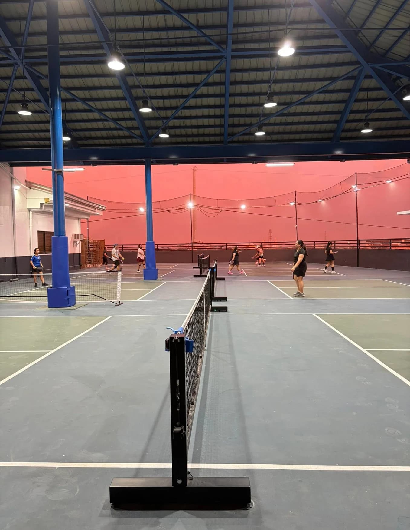 Players rally on a SkyCourt court while a vivid coral sunset glows beyond the rooftop.